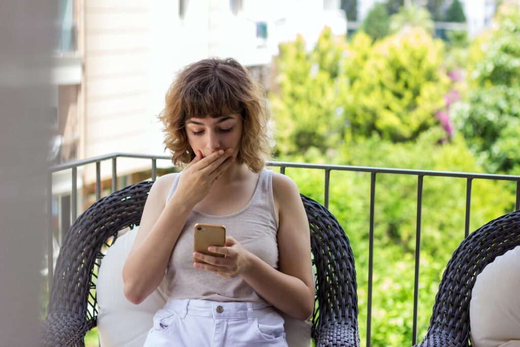 Pretty young girl with phone on apartment balcony. Video chatting with friends with shocking news